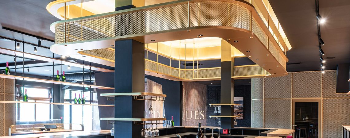 A contemporary wine tasting space features M22-83 brass woven wire mesh as a ceiling gantry that forms a sculptural canopy above the bar.
